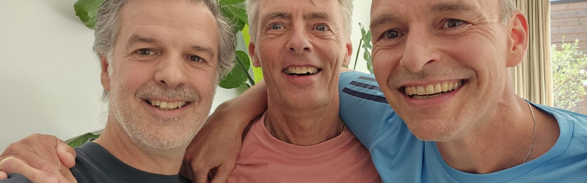 Drie mannen staan naast elkaar met armen over elkaars schouders. Ze kijken lachend in de camera. Ze zijn al wat ouder maar zeg dat maar niet tegen ze.