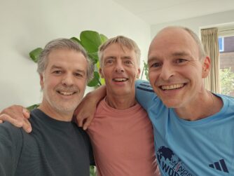 Drie mannen staan naast elkaar met armen over elkaars schouders. Ze kijken lachend in de camera. Ze zijn al wat ouder maar zeg dat maar niet tegen ze.