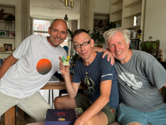 Drie mannen kijken blij in de camera. Dolf draagt een bril en houdt een gebakje beet met twee kaarsjes in de vorm van het getal 62.