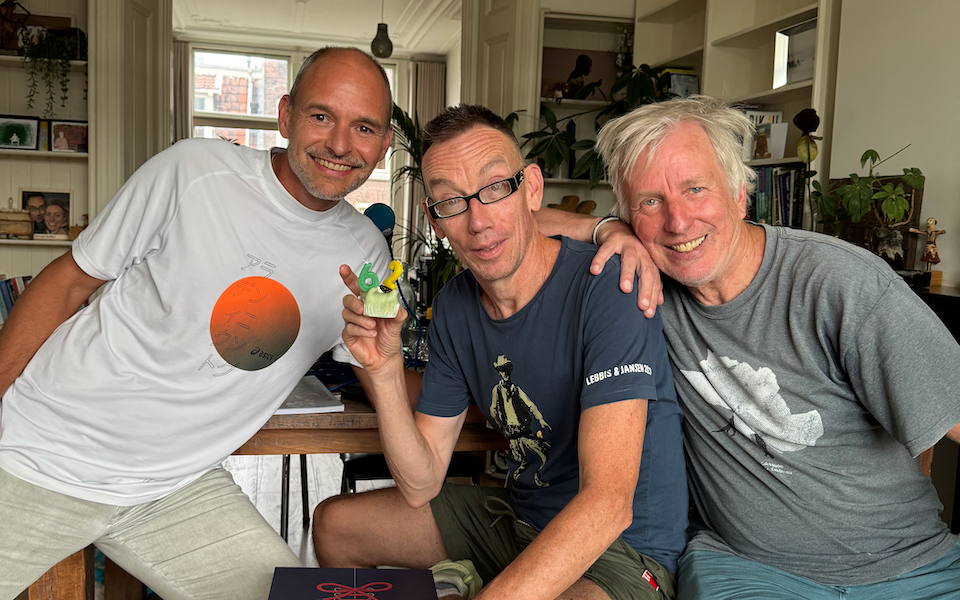 Drie mannen kijken blij in de camera. Dolf draagt een bril en houdt een gebakje beet met twee kaarsjes in de vorm van het getal 62.