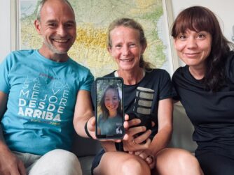 Jose kijkt lachend in de camera en houdt een telefoon beet waarop Noor te zien is. Naast Jose zit Irene in een korte hardloopshort en daarnaast zit Anne