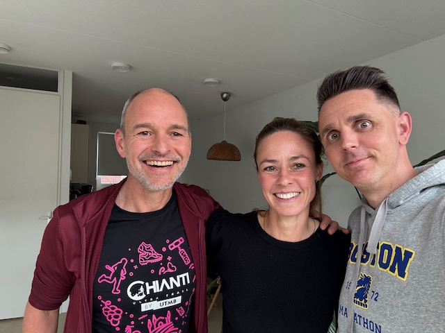 Jose staat breed lachend naast Hannah, een vrouw met een vrolijke lach, een open gezicht en lang donkerblond haar in een staart. Wouter krijgt vrolijk in de camera en draagt een grijze hoodie met het logo van de Boston marathon.